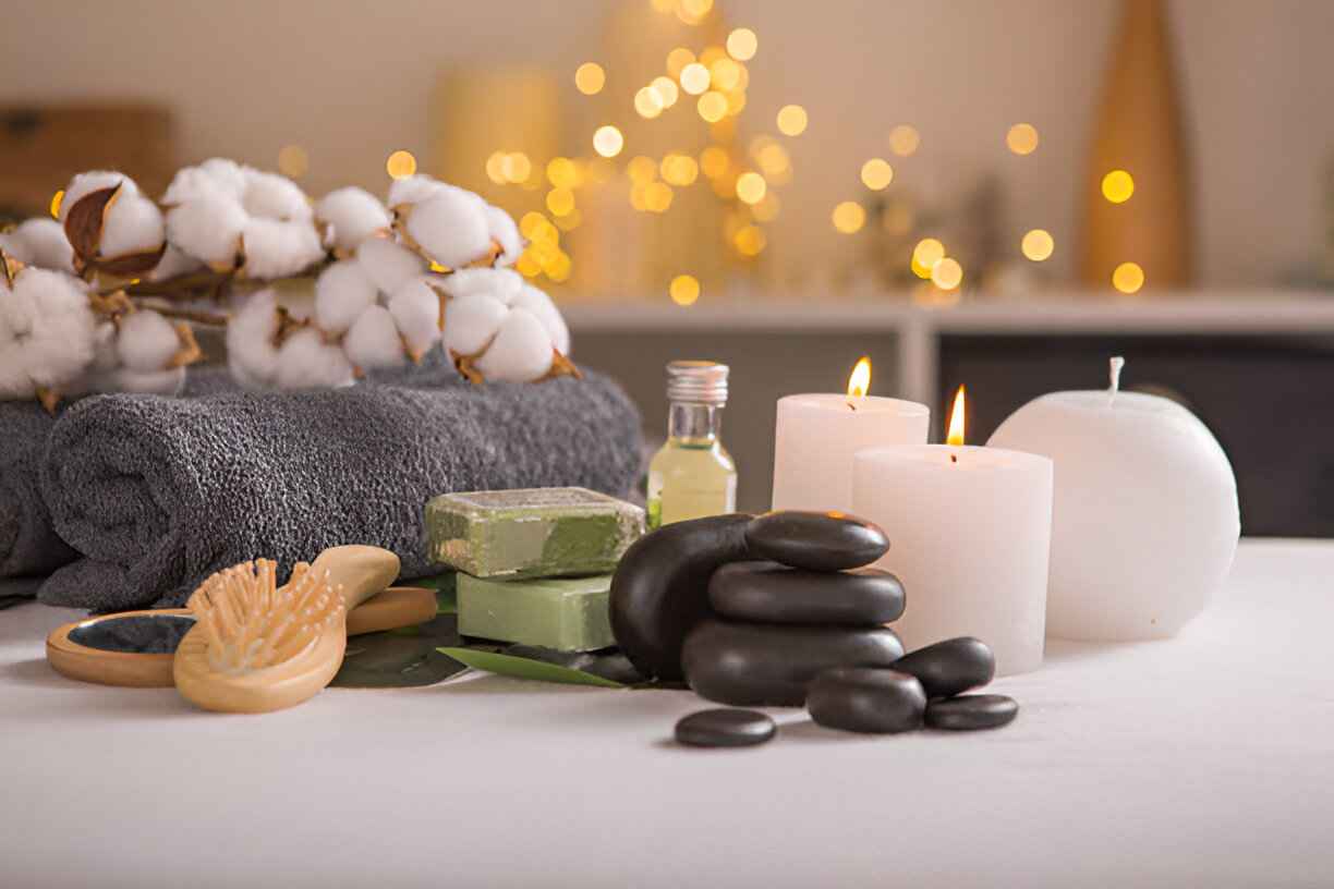 Spa setup with candles, stones, cotton flowers, towel, soap, and oil in a soft glowing background.